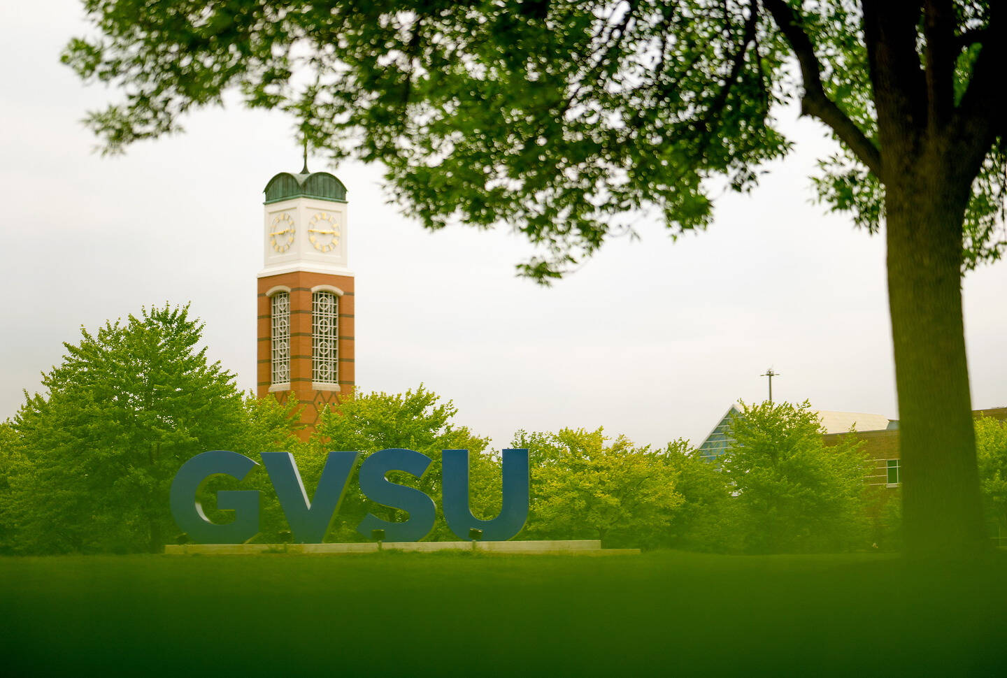 A clock tower stands behind large blue "GVSU" letters on a grassy field, framed by trees under an overcast sky.